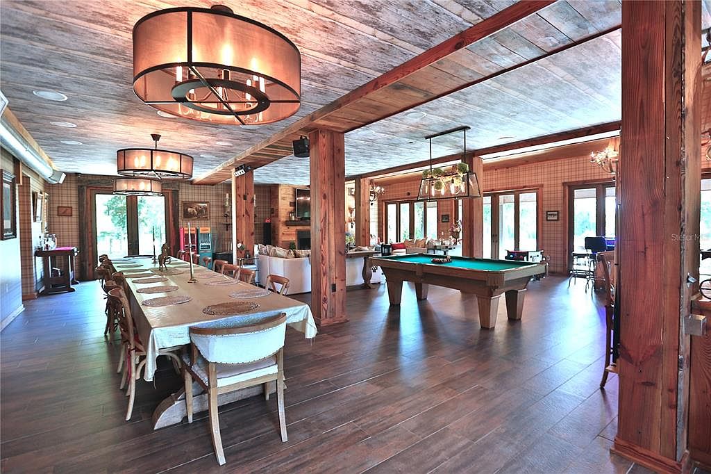 This expansive living room features a rustic yet elegant design with wood-paneled ceilings and support beams. The room is furnished with a long dining table, a pool table, and comfortable seating areas, creating a versatile space for both dining and recreation. Large windows provide ample natural light, enhancing the warm and inviting atmosphere.