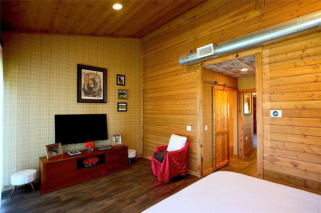 This is a cozy primary bedroom featuring wood-paneled walls and ceiling, creating a rustic and warm atmosphere. The room includes a TV area with a wooden console, a comfortable chair, and a glimpse into an adjacent hallway through a sliding barn door. The overall impression is inviting and comfortable, perfect for relaxation.