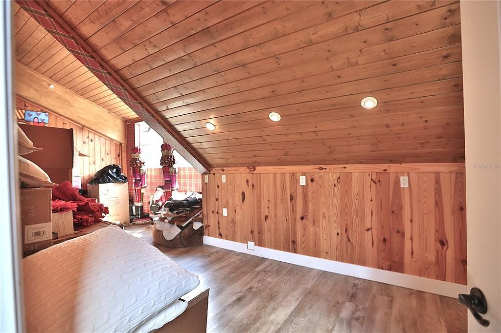 This is an interior shot of an attic space, featuring wood paneling on the walls and ceiling. The room is well-lit with recessed lighting and natural light from a window. The attic appears to be used for storage, with boxes and other items visible.