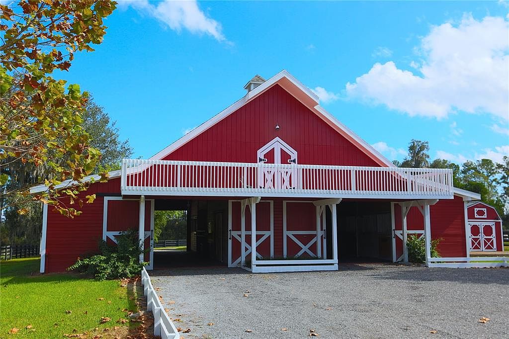 The image showcases the front view of a striking red barn with white trim, featuring a distinctive second-story balcony. The barn's design includes large doors and a well-maintained gravel driveway, suggesting a functional and aesthetically pleasing equestrian property. The clear blue sky and surrounding greenery enhance the property's appeal, creating a picturesque and inviting scene.