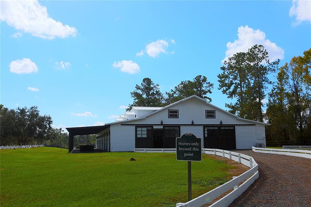 The image showcases the front exterior of a white barn-style building with black accents, set against a bright blue sky with scattered clouds. A well-manicured green lawn extends to the left, while a gravel driveway bordered by a white fence leads to the building on the right. A sign indicating 'Horses only beyond this point' is visible in the foreground, suggesting the property's equestrian use.