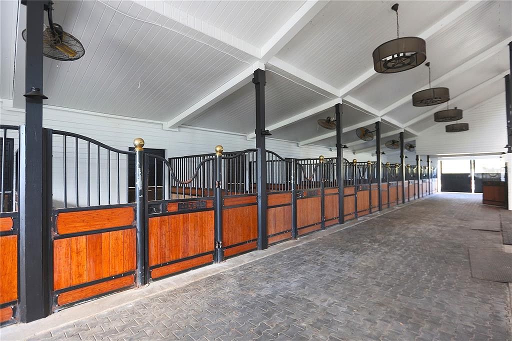 This image showcases the interior of a horse stable, featuring a row of stalls constructed with dark metal frames and warm-toned wood paneling. The stalls are lined along a paved walkway, and overhead, ceiling fans provide ventilation. The space is well-lit and appears clean and well-maintained, suggesting a high-quality equestrian facility.