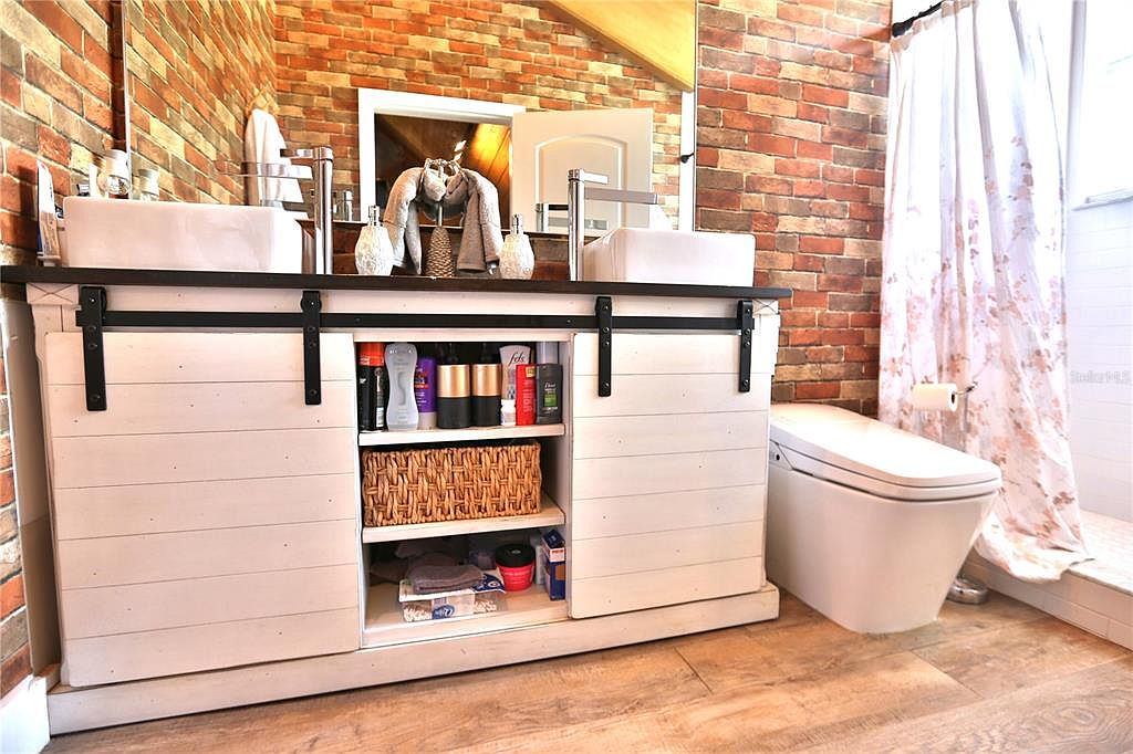 This is a well-lit primary bathroom featuring a double vanity with white vessel sinks and a dark countertop. The vanity has sliding barn doors revealing storage shelves. The walls are brick, adding a rustic touch, and a modern toilet is visible next to a shower with a floral curtain.