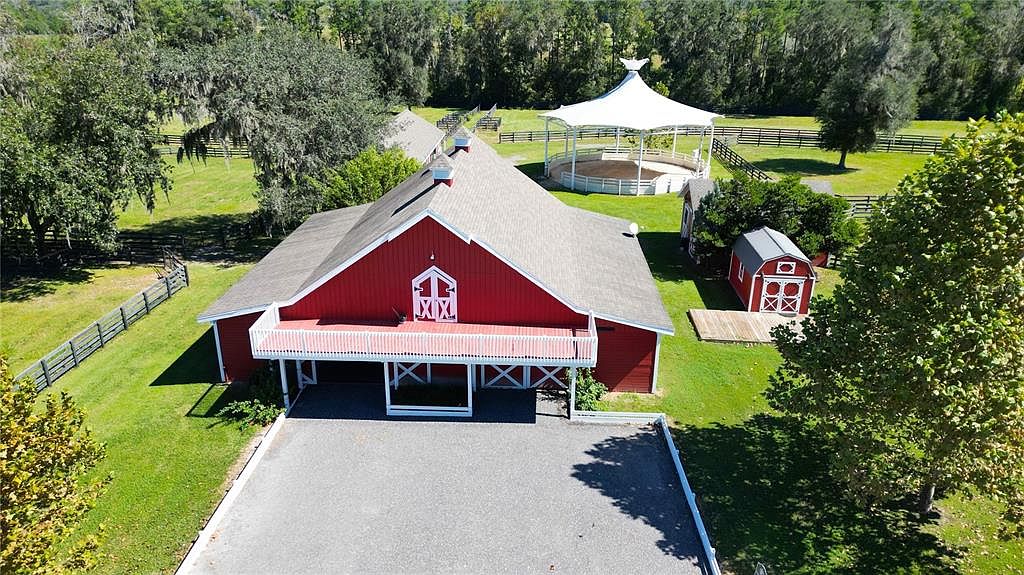 This aerial shot showcases a charming red barn-style house with a gray roof, complemented by a white balcony and matching trim. The property includes a separate red outbuilding and a white gazebo-like structure, all set within a lush green landscape with mature trees and fencing. A paved driveway leads up to the main building, enhancing the property's appeal.