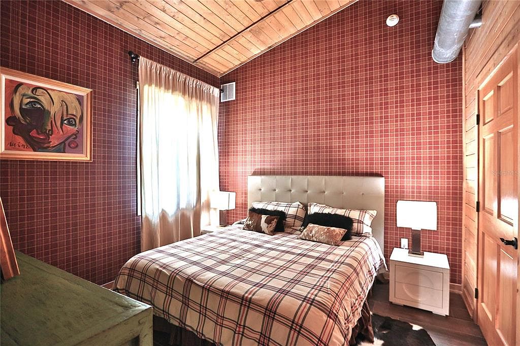 This is a cozy bedroom featuring red plaid wallpaper and a wooden ceiling. The room is furnished with a bed covered in a matching plaid comforter, a white nightstand with a lamp, and a framed painting on the wall. A window with light-colored curtains provides natural light, creating a warm and inviting atmosphere.