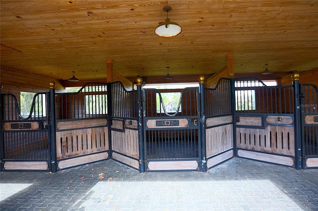 The image showcases the interior of a horse stall area, featuring wooden stalls with black metal bars and accents. The ceiling is made of wood planks, and lighting fixtures are visible. The floor is paved with interlocking bricks, creating a clean and functional space for horses.