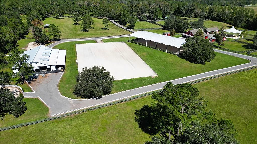 This aerial view showcases a sprawling equestrian estate. The property features a large riding arena, a covered training facility, stables, and a main residence with outbuildings, all surrounded by lush green pastures and mature trees. The layout emphasizes functionality and luxury, creating an impressive and well-maintained property.