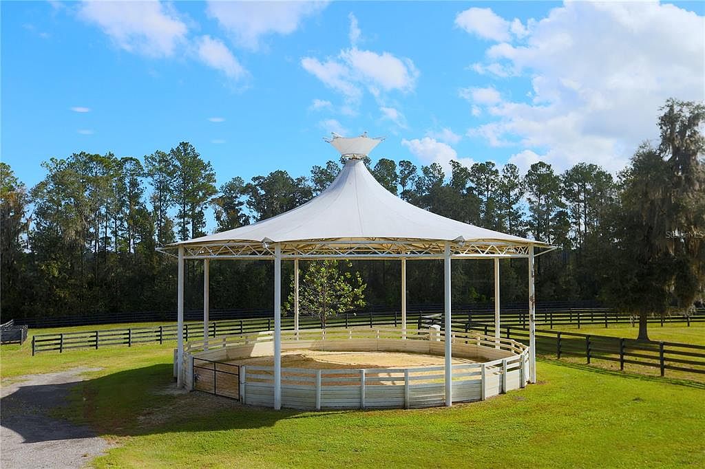 The image showcases a well-maintained outdoor riding arena with a white tent-like structure providing shade. The arena is surrounded by a white fence and lush green grass, with a backdrop of mature trees and a black wooden fence marking the property line. The scene conveys a sense of tranquility and upscale rural living.