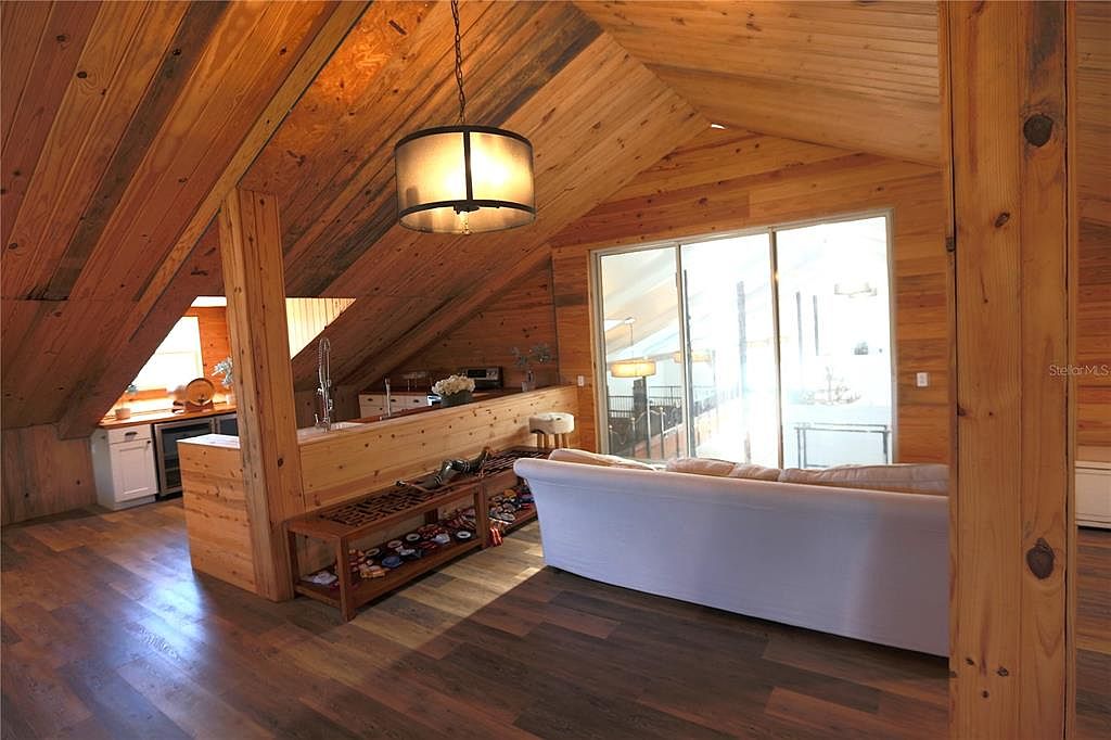 This interior shot showcases a cozy living room with a distinctive wood-paneled ceiling and walls, creating a warm, rustic ambiance. A modern white sofa sits in front of large sliding glass doors that flood the room with natural light. The open layout includes a partial view of a kitchen area, enhancing the sense of spaciousness.