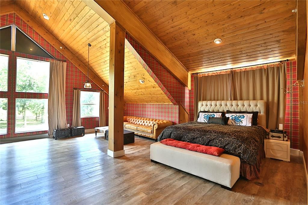 This is a primary bedroom featuring a unique design with wood-paneled ceilings and plaid wallpaper. The room includes a large bed with a tufted headboard, a leather sofa, and large windows that provide ample natural light. The overall style is rustic and cozy, creating a warm and inviting atmosphere.