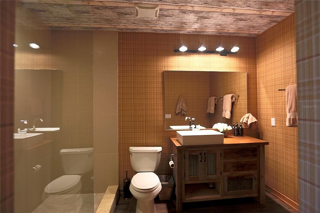 This is a well-lit bathroom featuring a unique design with a rustic touch. The walls are covered in a grid-patterned tile, complemented by a wooden ceiling. A modern sink sits atop a distressed wooden vanity, and the room includes a toilet and a separate shower area, creating a functional and stylish space.