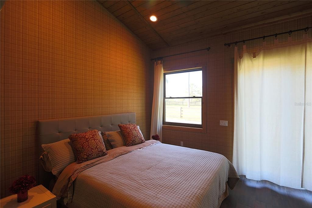 This is a cozy bedroom featuring a queen-sized bed with a tufted headboard and patterned throw pillows. The walls are covered in a textured, grid-like wallpaper, and the ceiling is wood paneled. Natural light streams in through a window, complemented by light-colored curtains, creating a warm and inviting atmosphere.