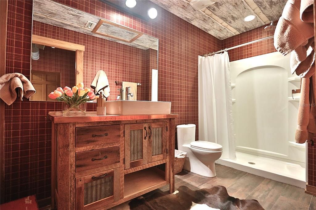 This is a rustic-style bathroom featuring a wooden vanity with a vessel sink, a large mirror, and a walk-in shower with a white curtain. The walls are covered in a red plaid pattern, and the ceiling has a distressed wood finish. The flooring appears to be wood-look tile, and a cowhide rug adds a touch of warmth to the space.