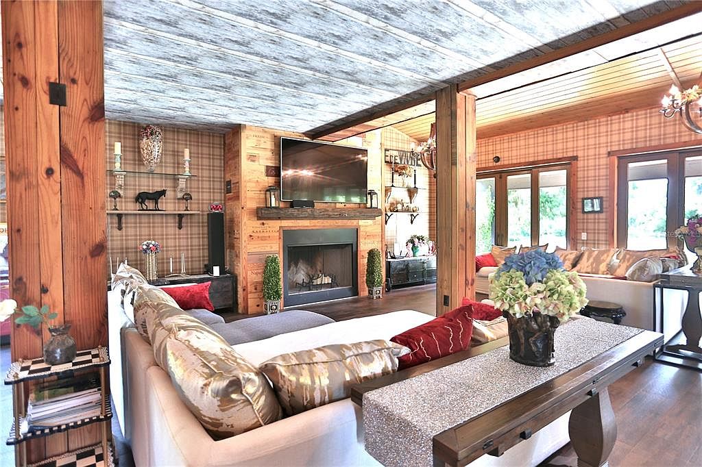 This is an interior shot of a living room featuring a rustic yet elegant design. The room includes a fireplace with a wooden surround, a large television mounted above the mantel, and comfortable seating arrangements. The walls are decorated with a plaid pattern, and the ceiling has a unique wooden plank design, creating a warm and inviting atmosphere.