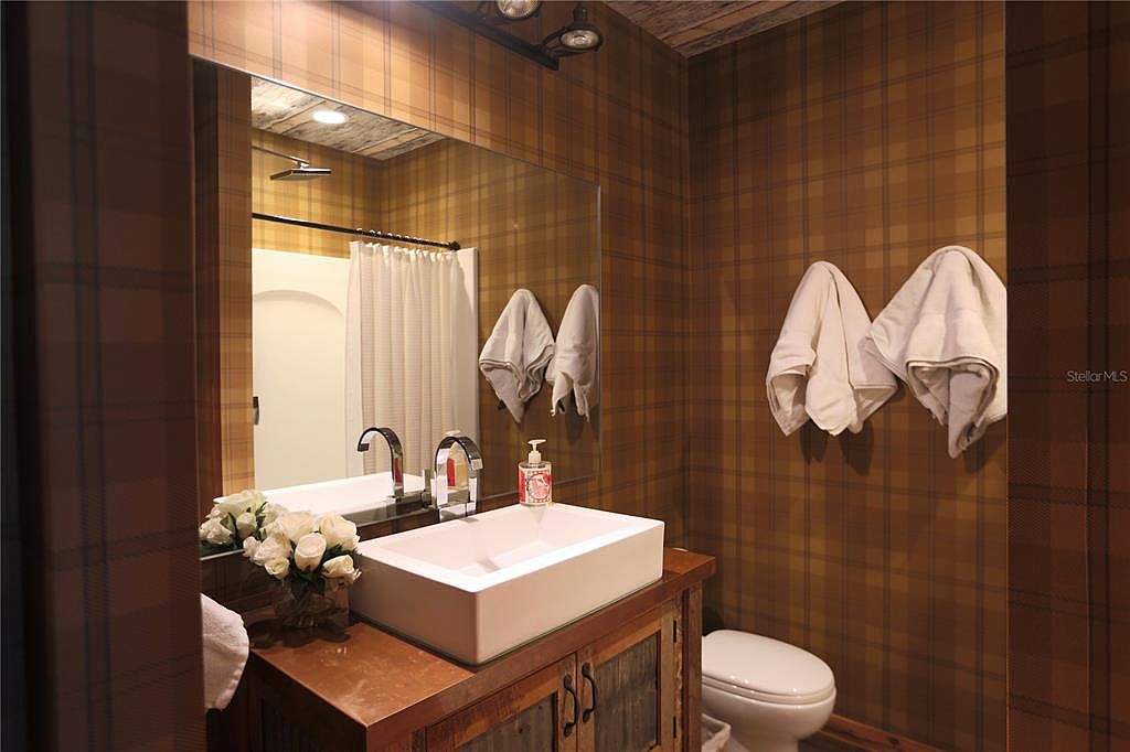 This is a cozy bathroom featuring a square white sink atop a rustic wooden vanity. The walls are covered in a brown plaid wallpaper, complemented by white towels and a toilet. The mirror reflects a shower with a white curtain, creating a warm and inviting space.