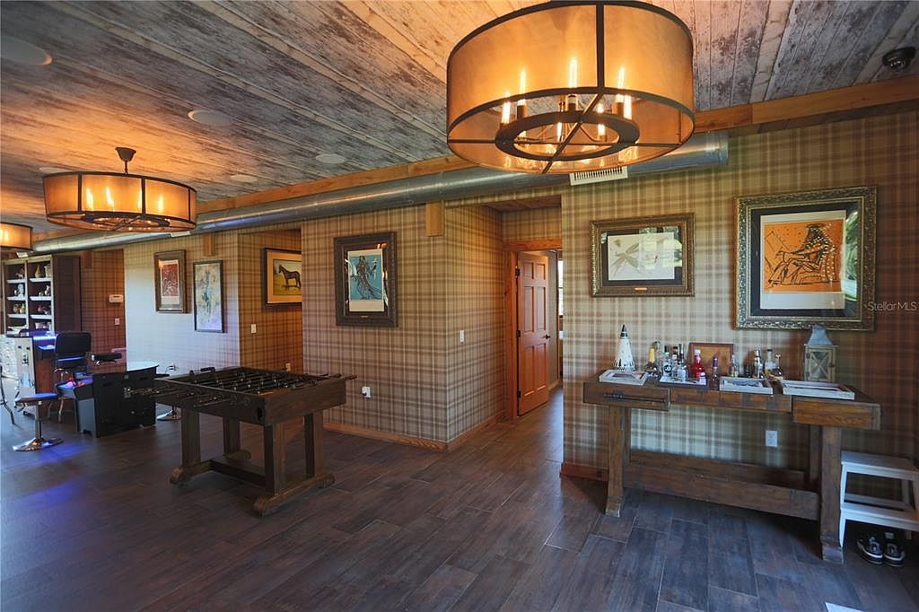 This interior shot showcases a rustic-style living room featuring wood-paneled walls and ceiling, complemented by dark wood flooring. The room is furnished with a foosball table and a console table displaying decorative items and glassware. Two large, round chandeliers provide warm lighting, enhancing the cozy and inviting atmosphere of the space.