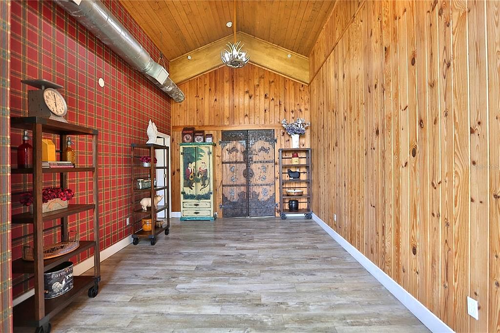 The image showcases an interior hallway with a unique blend of rustic and eclectic design elements. The walls feature a combination of red plaid wallpaper and natural wood paneling, complemented by light wood-look flooring. The hallway is furnished with decorative shelving units and antique-style doors, creating a visually interesting and inviting space.