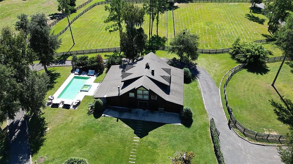 This aerial view showcases a stunning property featuring a large, dark-colored house with a complex roof design, a swimming pool with lounge chairs, and a long driveway leading to the house. The surrounding landscape includes expansive green lawns, fenced pastures, and mature trees, creating a sense of privacy and luxury. The image highlights the estate-like setting and the well-maintained grounds.