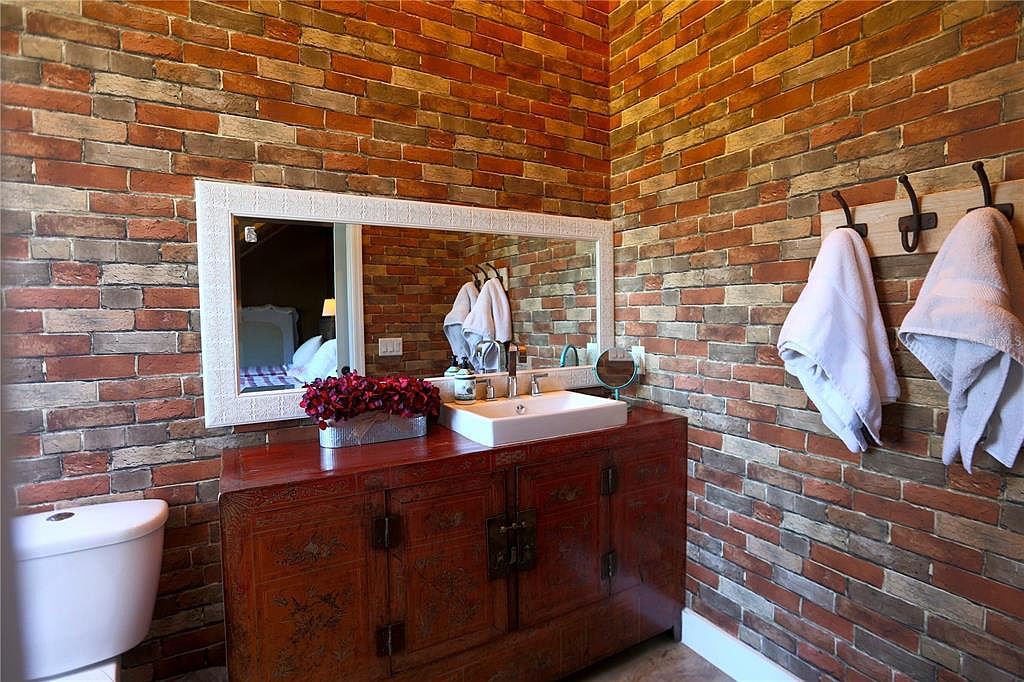 This bathroom features a unique brick wall design, complemented by a rustic wooden vanity with an ornate design. A white framed mirror hangs above the vanity, reflecting a glimpse into another room. Two white towels hang on hooks, adding a touch of simplicity to the space.