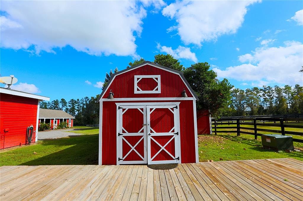 The image showcases a charming red barn with white trim, set against a backdrop of a bright blue sky with fluffy white clouds. A wooden deck extends from the barn, leading to a well-maintained lawn and a black wooden fence in the background. The scene evokes a sense of rustic charm and tranquility, ideal for a country estate.