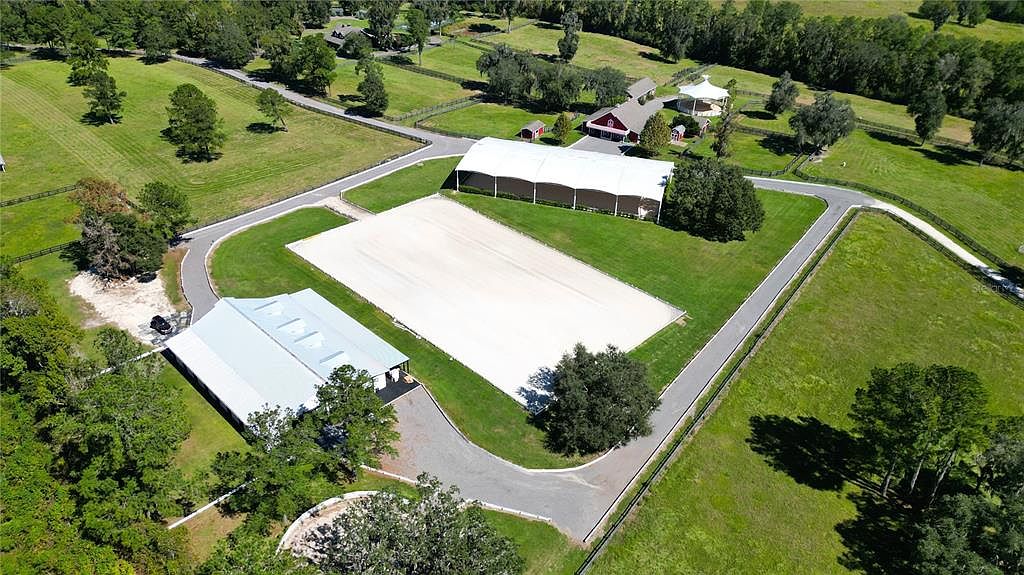 This aerial shot showcases a well-maintained equestrian estate. The property features a large outdoor riding arena, a covered arena, and a stable building, all surrounded by lush green pastures and mature trees. A winding driveway connects the various structures, creating a cohesive and appealing layout.