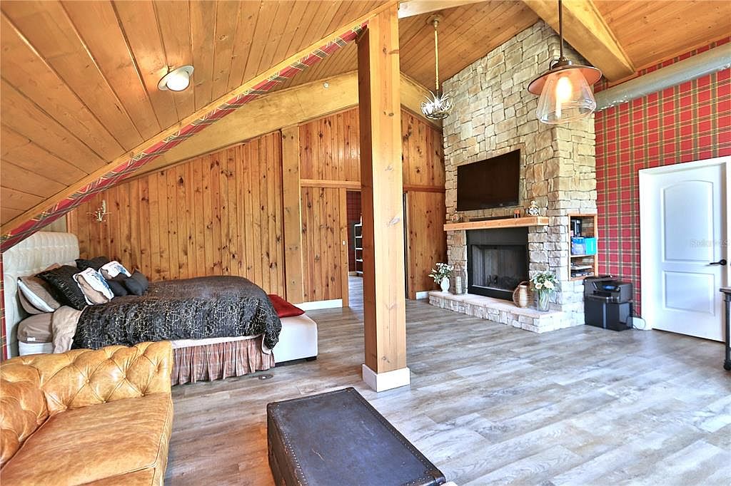 This is a spacious primary bedroom featuring wood-paneled walls and ceiling, a stone fireplace with a mounted TV, and a cozy bed. The room is decorated with a mix of textures and patterns, including plaid wallpaper and a tufted leather couch. The overall impression is warm and inviting, with a rustic yet comfortable feel.