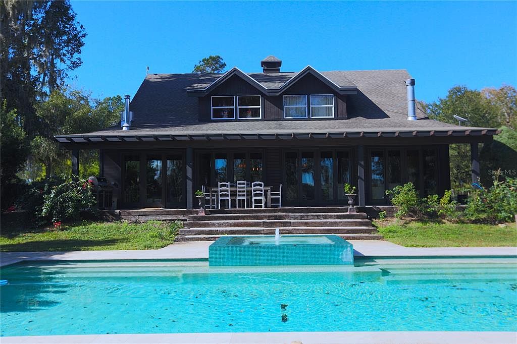 The image showcases a beautiful backyard featuring a large swimming pool with a modern, tiled fountain feature. The pool is surrounded by well-maintained grass and leads up to a house with a covered patio and multiple glass doors. The overall impression is one of luxury and relaxation, perfect for outdoor entertaining.