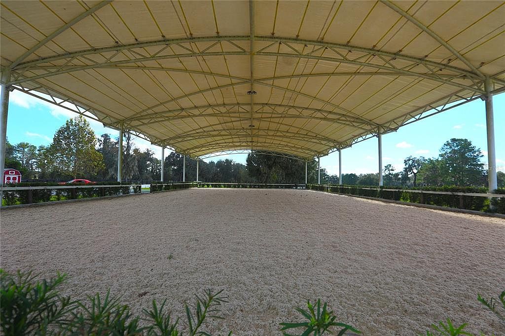The image showcases a large, covered equestrian arena with a sand surface, surrounded by manicured hedges and greenery. The structure features a light-colored canopy supported by white pillars, creating an open and airy space. In the background, trees and a glimpse of a red building add depth to the scene.