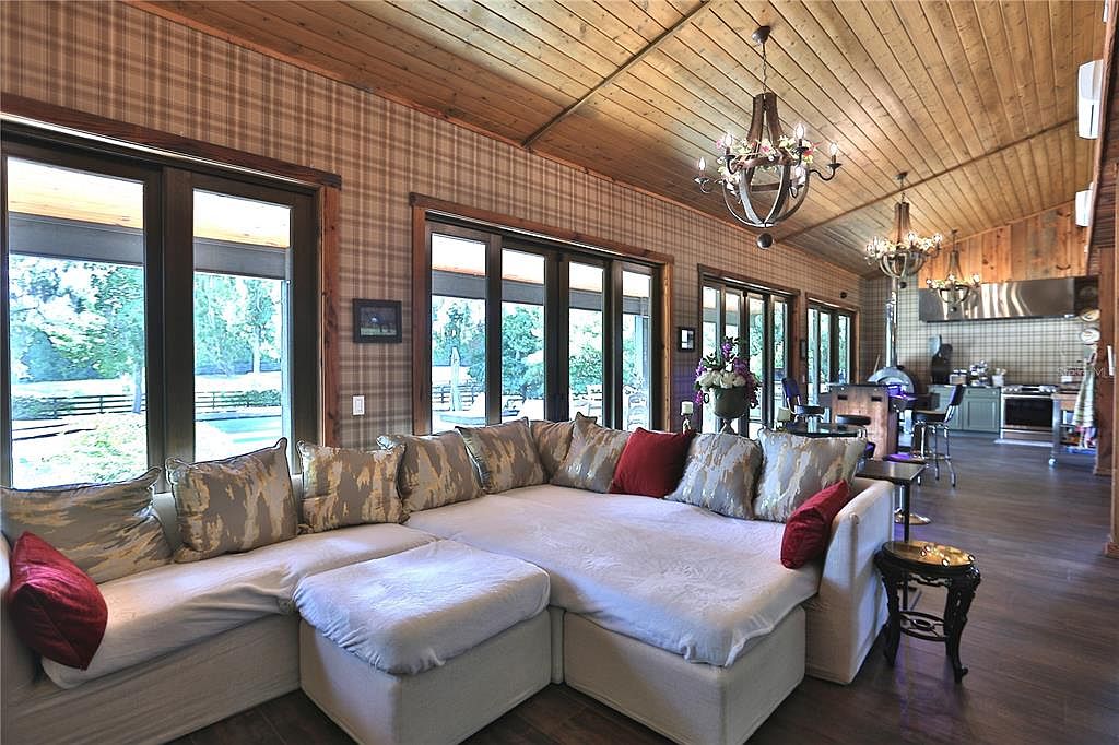 This interior shot showcases a spacious living room with a large, sectional sofa adorned with decorative pillows. The room features a unique blend of rustic and elegant design elements, including wood-paneled ceilings, plaid wallpaper, and a decorative chandelier. Large windows provide ample natural light and views of the exterior landscape, while the open floor plan seamlessly connects the living area to the kitchen.