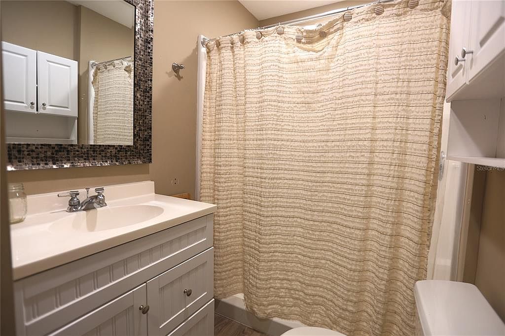 This is a bathroom featuring a white vanity with a white countertop and a decorative mirror. A beige shower curtain with horizontal stripes covers the shower/tub area. A toilet is visible to the right of the shower, and the walls are painted in a neutral tone, creating a clean and functional space.