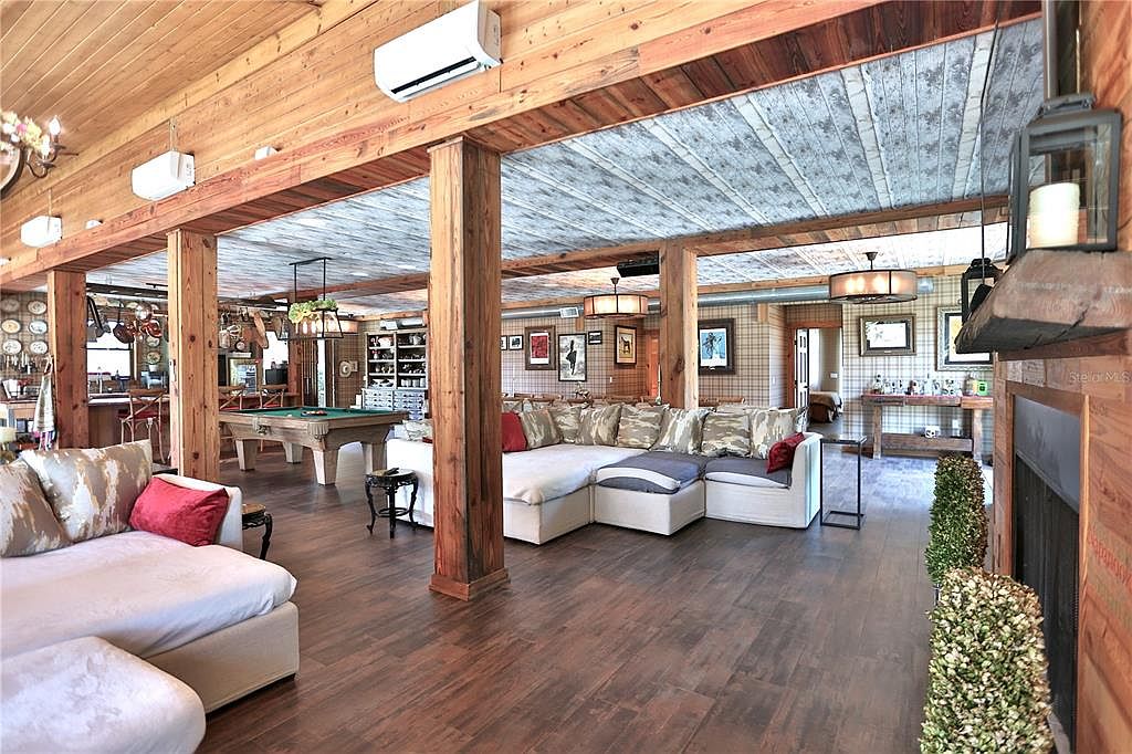 This is an interior shot of a spacious living room featuring wooden floors, exposed wooden beams, and a unique ceiling design. The room is divided into sections with large wooden pillars, creating distinct areas for seating and recreation, including a pool table. The overall style is rustic and inviting, with a focus on natural materials and a comfortable atmosphere.