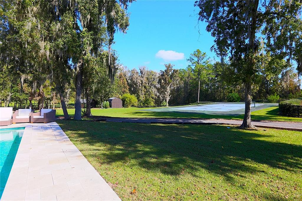 The image showcases a well-maintained yard with lush green grass, mature trees, and a glimpse of a tennis court in the background. A pool is visible on the left side of the image, along with patio furniture. The scene evokes a sense of tranquility and spaciousness, highlighting the property's outdoor amenities.