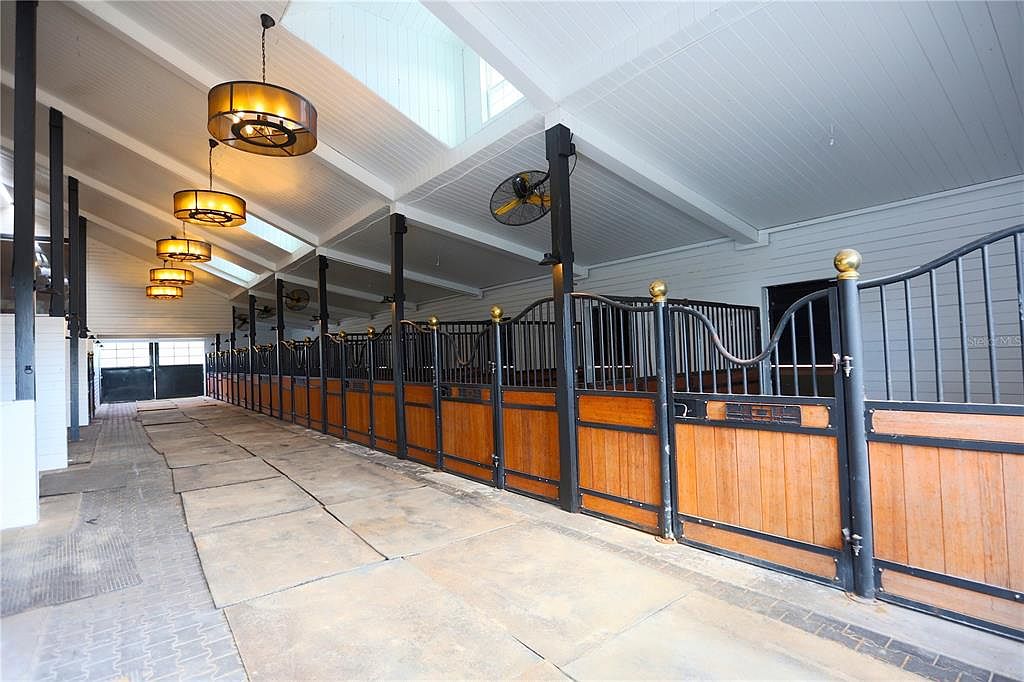 This image showcases the interior of a horse stable, featuring a long corridor lined with individual stalls. The stalls are constructed with wood and black metal, topped with decorative gold accents. Overhead, modern light fixtures and ceiling fans provide illumination and ventilation, while skylights allow natural light to filter in, creating a clean and well-maintained environment.