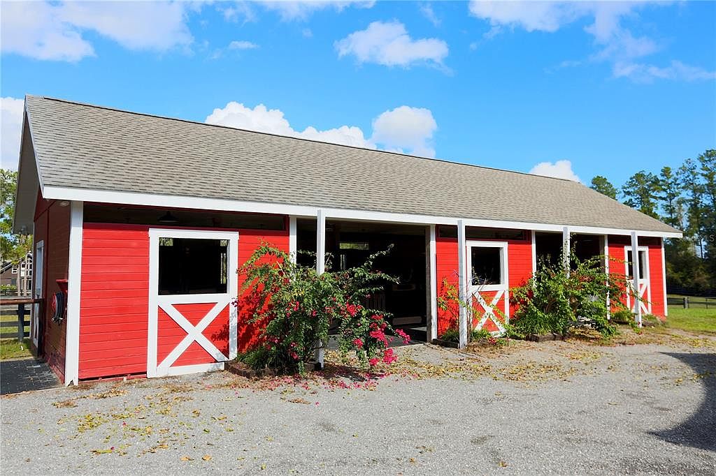The image showcases the front view of a charming red barn-style building with white trim and a gray roof. The structure features sliding barn doors and decorative 'X' details on the windows. Lush greenery with red flowers adds a touch of color to the gravel driveway, creating a welcoming and rustic aesthetic.