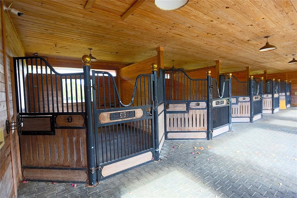 The image showcases a series of horse stalls inside a barn, constructed with wood and black metal. The stalls feature ornate designs and individual lighting fixtures, creating a luxurious and well-maintained environment for horses. The flooring is paved with gray bricks, and the overall impression is one of quality and care.