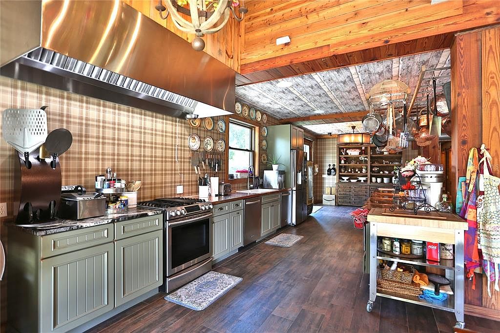 This is a warm and inviting kitchen with a rustic, country-style design. It features green cabinetry, stainless steel appliances, and a large stainless steel range hood. The walls are covered in a plaid wallpaper, and the ceiling is wood paneled, creating a cozy atmosphere. The kitchen also includes a mobile kitchen island with storage and a variety of decorative plates and cookware displayed on the walls and hanging from the ceiling.