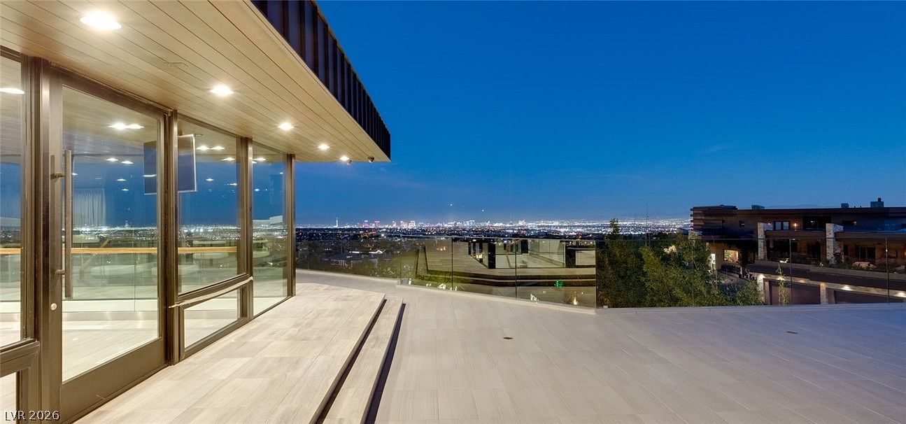 This expansive, modern terrace features light-toned tile flooring and a sleek glass railing that provides an unobstructed view of the city skyline at dusk. The space is accessed through large, floor-to-ceiling glass doors, with recessed lighting integrated into the deep roof overhang. The elevated perspective highlights the property's prime location and sophisticated architectural design.