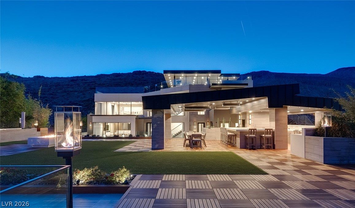 This contemporary luxury residence features a multi-level architectural design with expansive glass walls and a covered outdoor living space. The rear yard includes a manicured lawn, a patterned tile patio, and a built-in outdoor kitchen with bar seating, all set against a dramatic mountain backdrop at dusk. The scene is illuminated by warm accent lighting, creating an inviting and sophisticated atmosphere for evening entertaining.