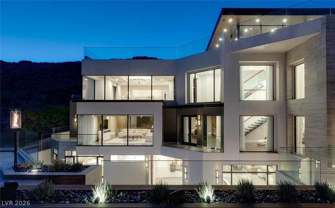 This contemporary multi-level residence features a striking architectural design with clean lines and extensive glass walls that offer a seamless connection between indoor and outdoor living spaces. The exterior is illuminated at dusk, highlighting the terraced levels, glass railings, and a landscaped foreground with decorative stone and lighting. The perspective captures the expansive rear facade, showcasing the modern aesthetic and the home's integration into the hillside setting.