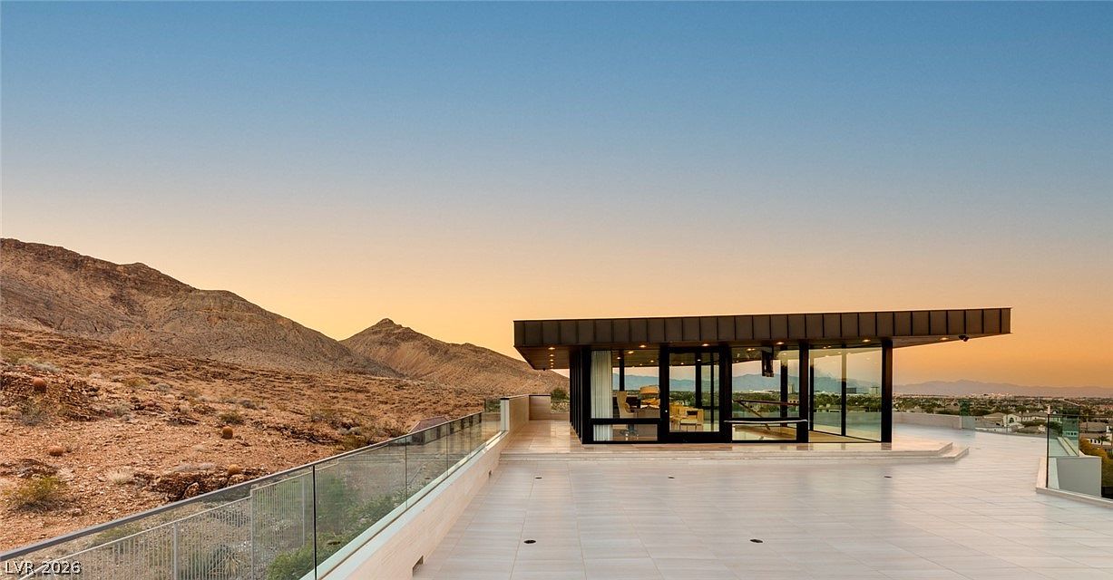 This expansive, modern terrace features light-toned tile flooring and a sleek glass railing that overlooks a desert landscape and distant city views. A contemporary structure with floor-to-ceiling glass walls sits on the deck, providing a seamless indoor-outdoor transition. The scene is captured at sunset, with the warm, golden light highlighting the rugged mountain backdrop and the clean, minimalist lines of the architecture.