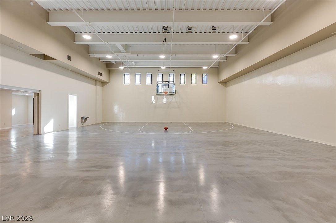This spacious indoor basketball court features a polished, light-colored concrete floor and high, exposed-beam ceilings. A basketball hoop is mounted on the far wall, centered beneath a row of rectangular windows, with a single basketball resting in the middle of the court. The room is painted in a neutral, light tone, creating an open and versatile recreational space.