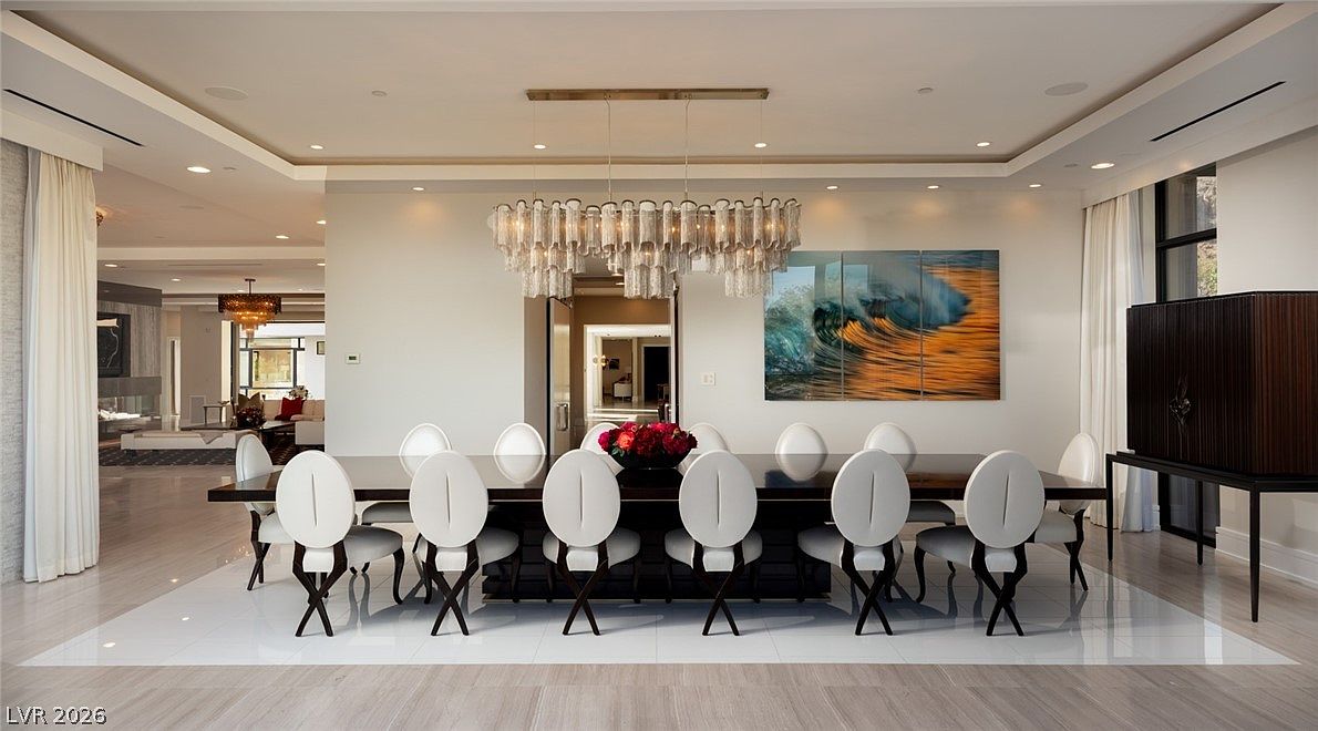 This elegant dining room features a long, dark-toned table surrounded by ten modern, oval-backed chairs with white upholstery and dark legs. A striking, multi-tiered crystal chandelier hangs above the table, serving as the room's focal point, while a large, vibrant wave-themed artwork adorns the wall. The space is finished with polished light-colored flooring and opens into an adjacent living area, creating an airy and sophisticated atmosphere.