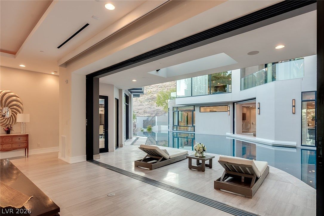 This contemporary living space features a seamless transition to an outdoor pool area through expansive, retractable glass walls. The interior is finished with light-toned flooring and minimalist decor, while the exterior showcases a modern pool deck with lounge chairs and a view of a rocky hillside. The open-concept design creates a bright, airy atmosphere that emphasizes indoor-outdoor living.