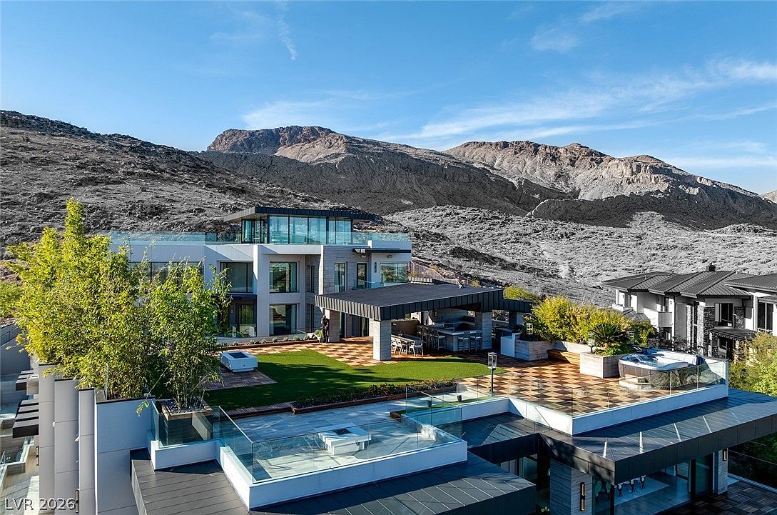 This aerial perspective captures a luxurious modern residence set against a dramatic, rugged mountain backdrop. The multi-level home features expansive terraces with checkered tile flooring, a private hot tub, and a covered outdoor kitchen area, all integrated with sleek glass railings. The architecture emphasizes clean lines and large windows, creating a seamless transition between the sophisticated indoor living spaces and the meticulously landscaped outdoor environment.