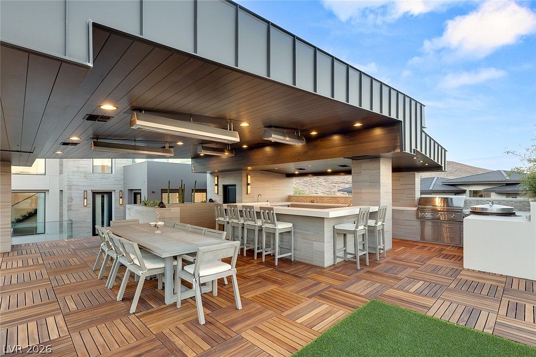 This expansive outdoor terrace features a covered dining and kitchen area with a modern, minimalist aesthetic. The space is equipped with a built-in grill, a stone-topped bar with seating, and a large dining table, all set upon a rich wood-tiled deck. The architectural design incorporates clean lines, recessed lighting, and overhead heaters, creating a sophisticated environment for outdoor entertaining.