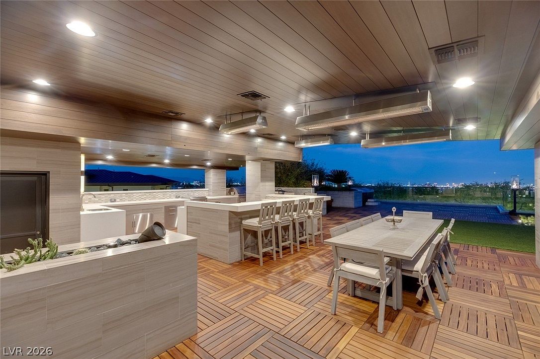 This expansive covered outdoor living space features a sophisticated kitchen area with a built-in grill and bar seating, complemented by a large dining table for alfresco meals. The area is finished with patterned wood decking and recessed ceiling lighting, creating a warm, inviting atmosphere that overlooks a distant city skyline. The perspective captures the seamless transition between the sheltered lounge area and the open-air deck, emphasizing a luxurious lifestyle perfect for entertaining.