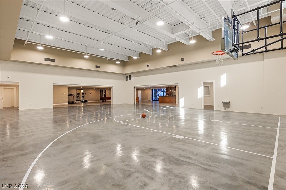 This expansive indoor sports court features a polished, reflective concrete floor marked with basketball court lines. The high, industrial-style ceiling is equipped with multiple pendant lights, and a basketball hoop is mounted on the right wall. Beyond the main court area, an open transition leads to a secondary space containing fitness equipment and a bar area, creating a versatile recreational environment.