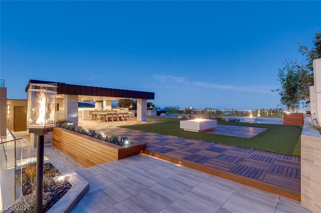 This expansive outdoor terrace features a sophisticated blend of wood decking and large-format stone tiles, creating a modern multi-level living space. The area is anchored by a covered dining pavilion with a long table, complemented by a central fire feature and a sleek glass-enclosed fire element in the foreground. The elevated perspective captures a panoramic view of the city skyline at dusk, emphasizing a luxurious and serene atmosphere perfect for evening entertaining.