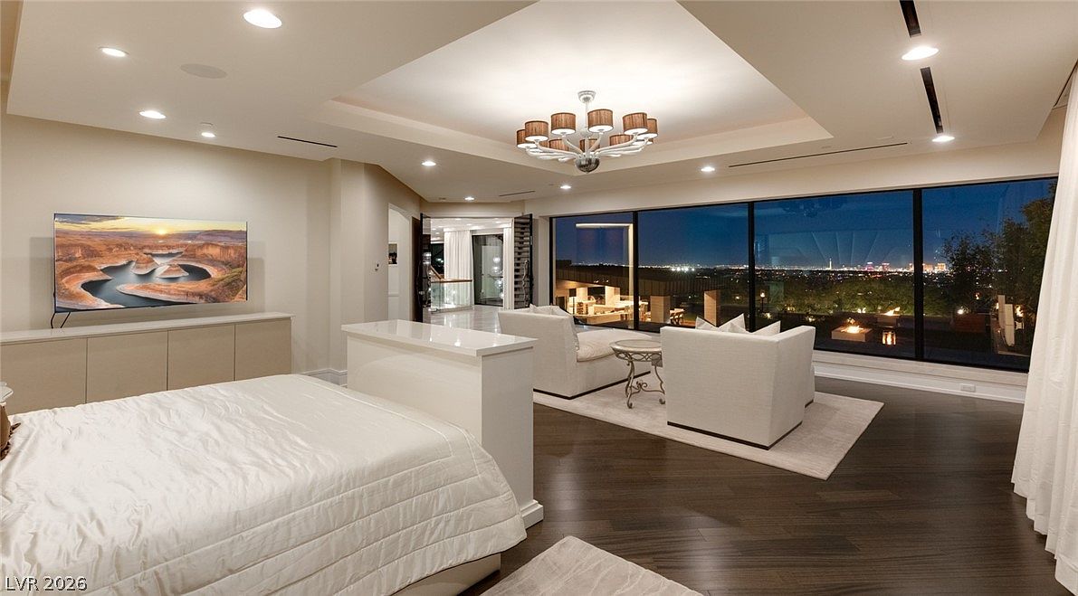 This expansive primary bedroom features a contemporary design with a neutral color palette and dark wood flooring. A large bed is positioned in the center of the room, separated from a cozy seating area by a low partition wall, while floor-to-ceiling windows offer a panoramic view of the city lights at night. The space is illuminated by a modern chandelier and recessed lighting, creating a sophisticated and tranquil atmosphere.