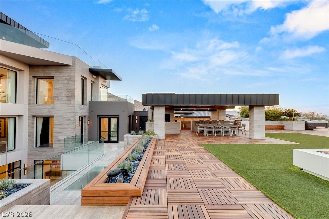 This expansive modern terrace features a combination of wood-tiled decking and manicured artificial turf, creating a sophisticated outdoor living space. A covered dining area with a built-in kitchen setup provides a shaded retreat, while a long, linear planter box filled with succulents adds a touch of greenery. The scene is framed by contemporary architecture with glass railings and large windows, offering a seamless transition between indoor and outdoor environments.